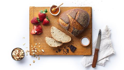 Fresh baked loaf of bread with seeds and grains on top. Two slices fanned in front of it with strawberries, honey and chocolate all on top of a cutting board.