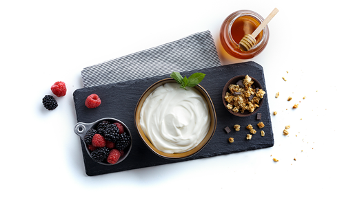 Fresh and smooth yogurt in a bowl with granola and berries in ramekins on both sides all sat on a slate tablet on a white background.