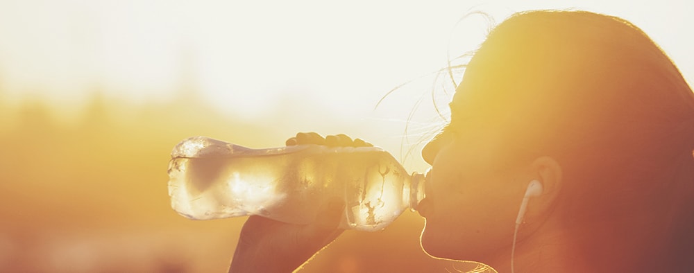 Woman with earbuds in sunlight drinking bottled water.