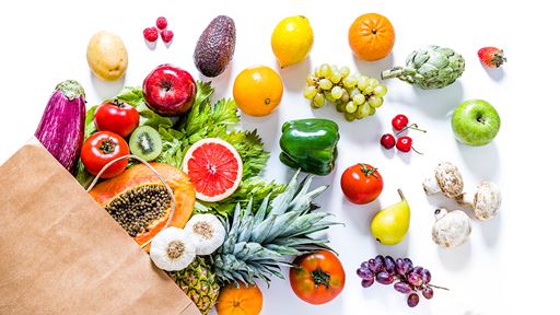 Brown grocery bag overflowing with a wide range of fruits and vegetables laid out on a white background.
