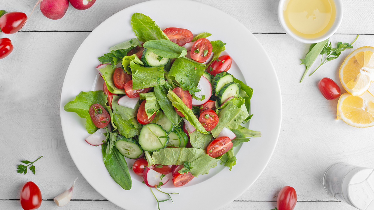 A simple salad with various greens and tomatoes and cucumber in a shallow circular white dish surrounded by other various vegetables.