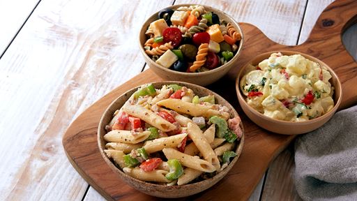 Three prepared versions of pasta salad in circular bowls atop a dark wood cutting board.