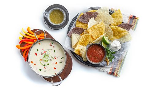A selection of various colored thin and crispy tortilla chips served with two different ramekins of salsa and a large side of white queso cheese dip.