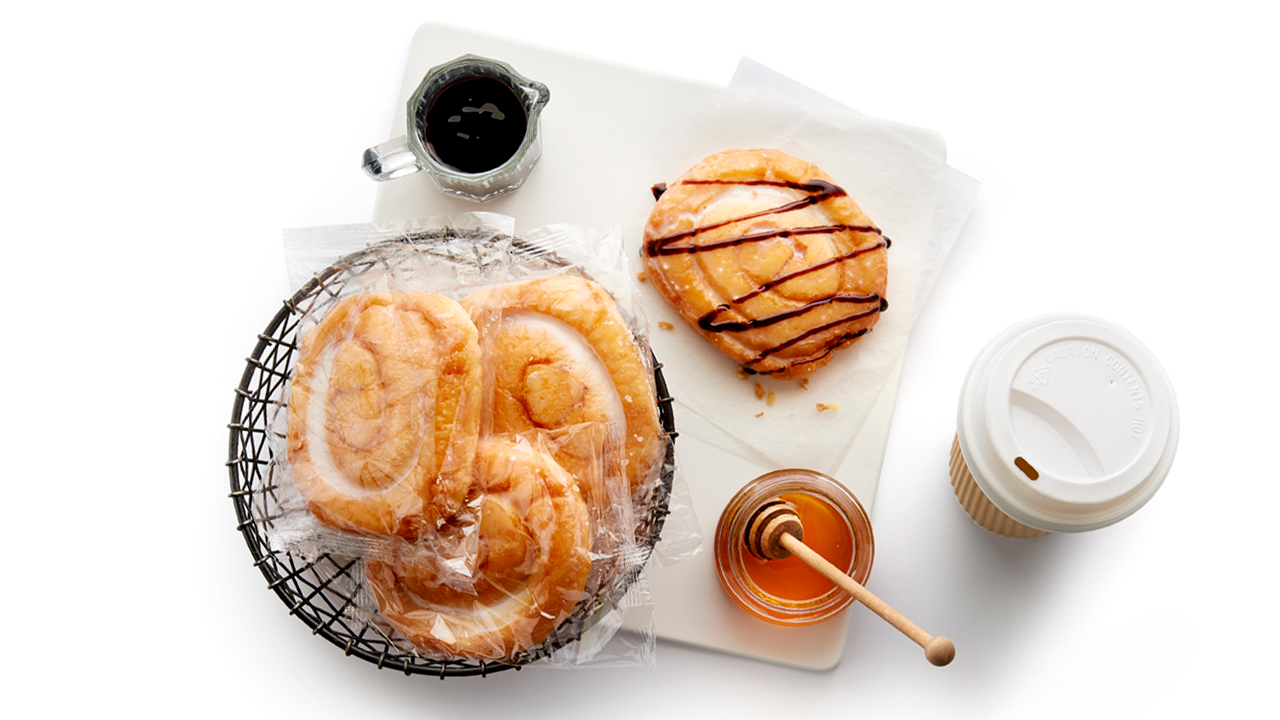 Packaged honey buns in bowl next to opened product with chocolate drizzle all atop of a marble served with fresh coffee.