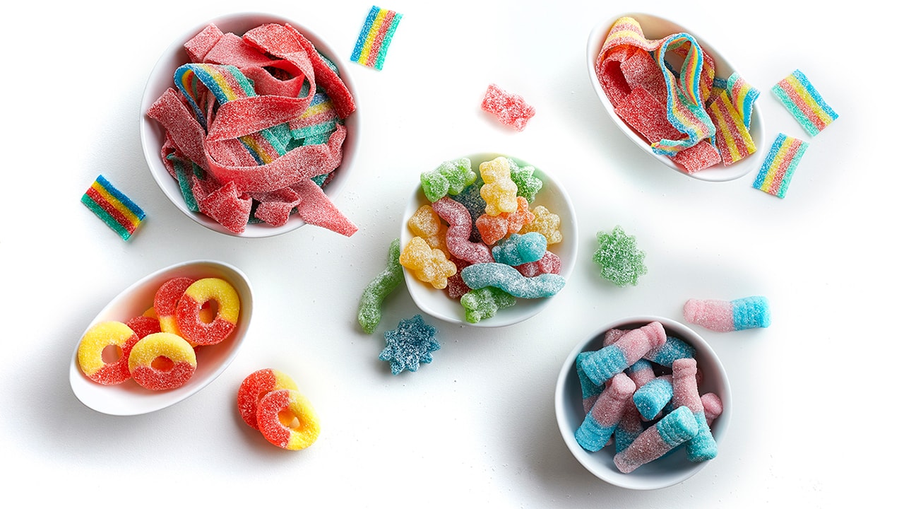 Five oval and round shaped white ramekins filled with various versions of sour and sanded candies.