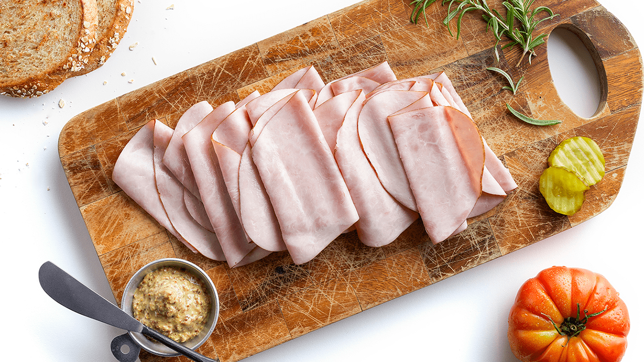 Sliced deli meat, mustard, pickles, bread, tomato on wooden cutting board.