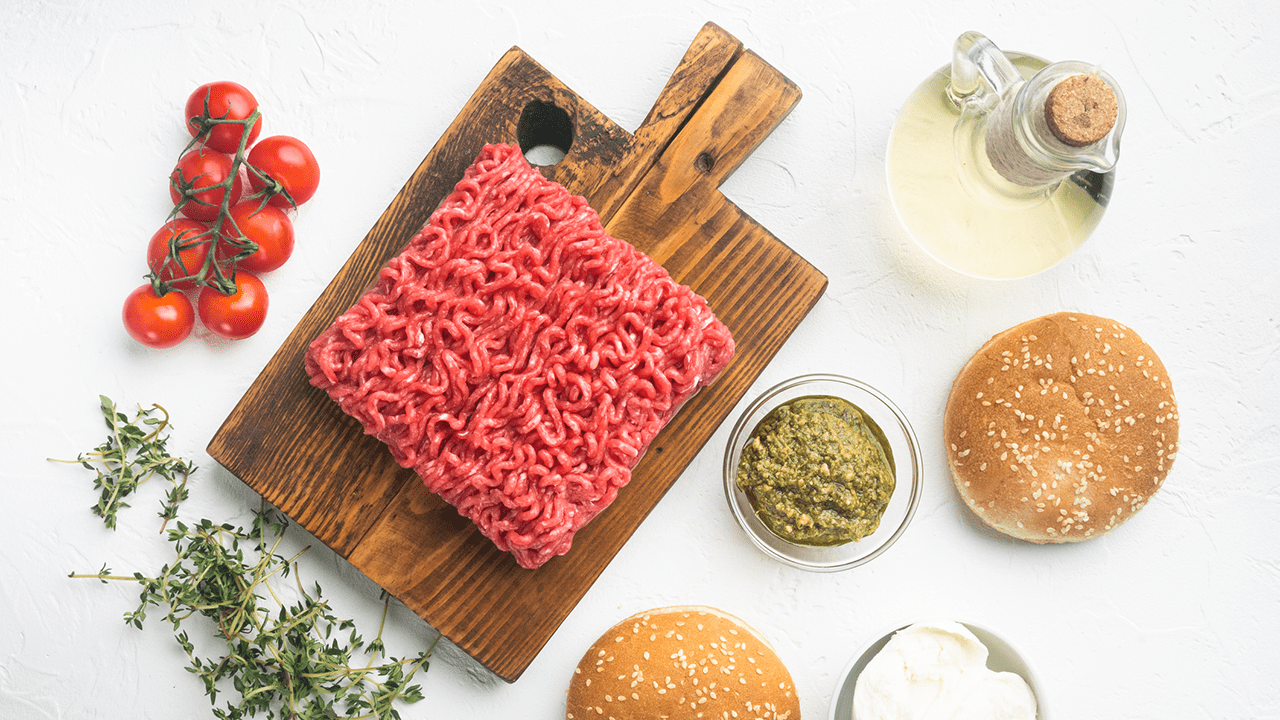 Ground beef on cutting board surrounded by bread and condiments.