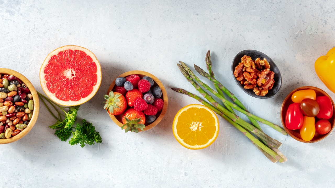 An organized and evenly spaced set of fruit and vegetables laid horizontally in a row on a white background.