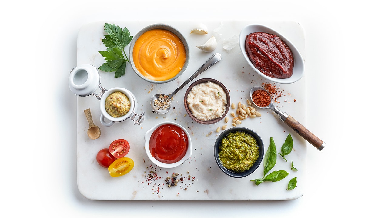 A white granite square slab topped with ramekins, containers, and spoons filled with 8 different sauces and dressings surrounded by spices, seasoning sand other ingredients.